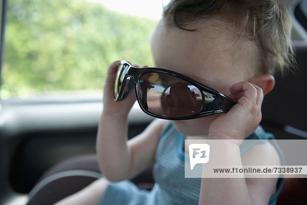 Ein kleiner Junge sitzt in einem stationären Auto und spielt mit einer Sonnenbrille.