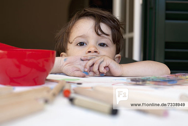 Ein Kind  das sich auf einen Tisch lehnt  der mit Buntstiften und Kunstzubehör übersät ist.