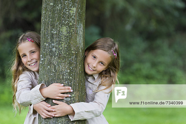 Zwillinge  Mädchen  9 Jahre  schauen links und rechts an einem Baum vorbei