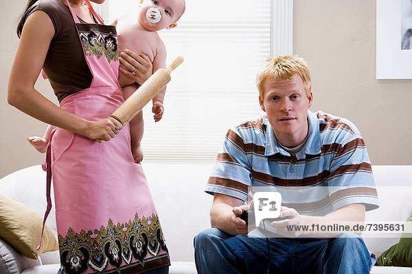 Man on sofa with video game controller and woman in apron with rolling pin holding baby