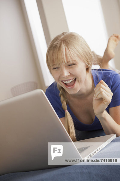 Laughing young woman lying on bed using laptop