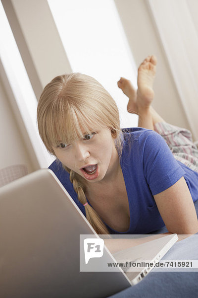 Surprised young woman lying on bed using laptop