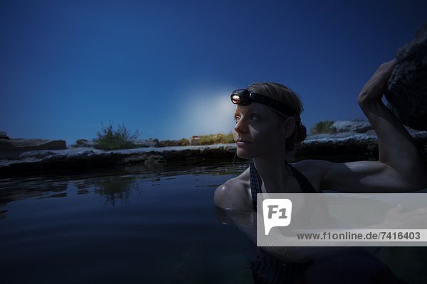 A girl sits in a natural hot springs with a headlight on.