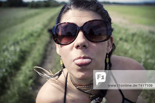 A young woman wearing sunglasses sticks her tongue out at the camera on a warm summer day.