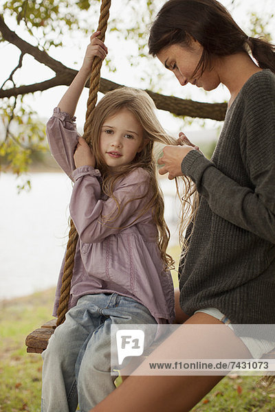 Mutter flechtet die Haare der Tochter auf einer Schaukel am Seeufer.