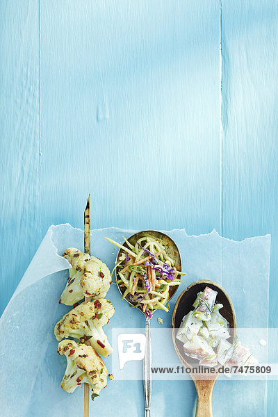 Overhead View of Grilled Cauliflower on Screwer and Spoons Filled with Salad on Wax Paper on Blue Wooden Table in Studio