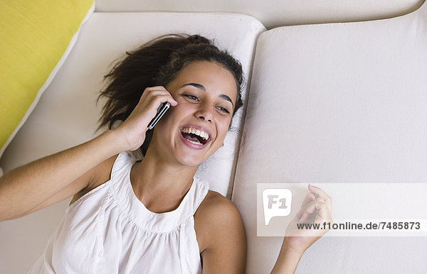 Teenager-Mädchen auf weißer Couch liegend und mit Handy  lächelnd