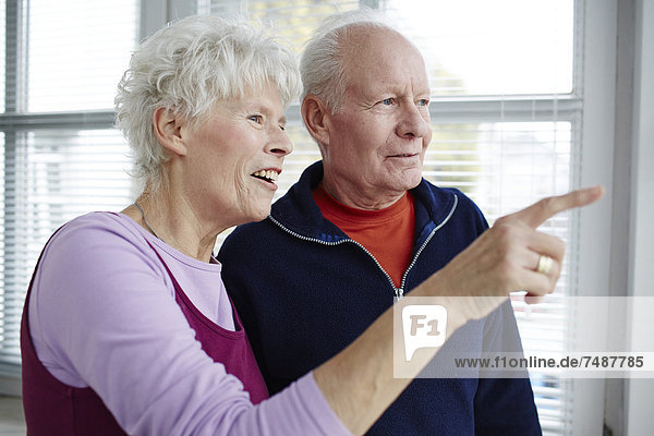 Deutschland  Düsseldorf  Seniorenpaar schaut durchs Fenster  lächelnd
