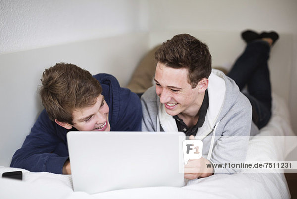 Happy young boys using laptop together while lying in bed