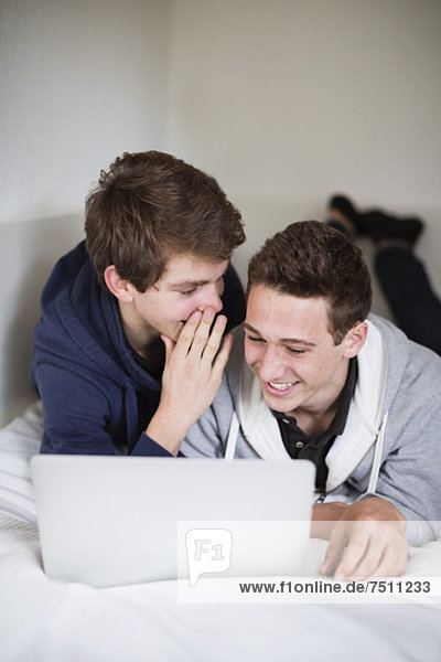 Young boy sharing secret with friend using laptop while lying in bed