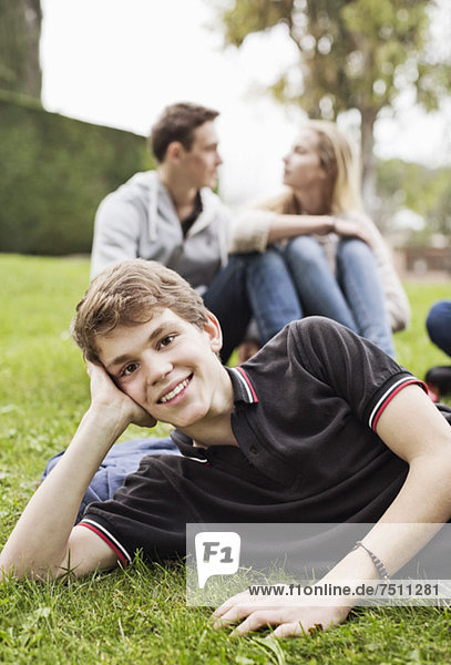 Porträt eines Jungen auf Gras liegend mit einem im Hintergrund sitzenden Paar