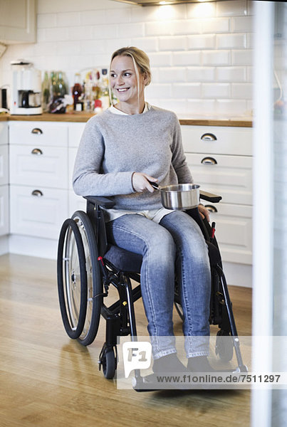 Glückliche behinderte Frau im Rollstuhl schaut weg  während sie den Topf in der Küche hält.