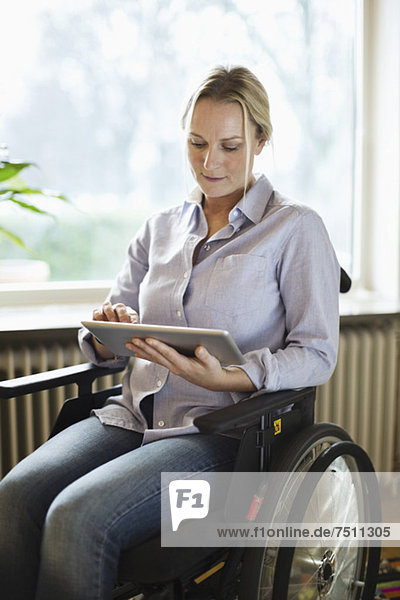 Behinderte Frau  die zu Hause im Rollstuhl sitzt und ein digitales Tablett benutzt