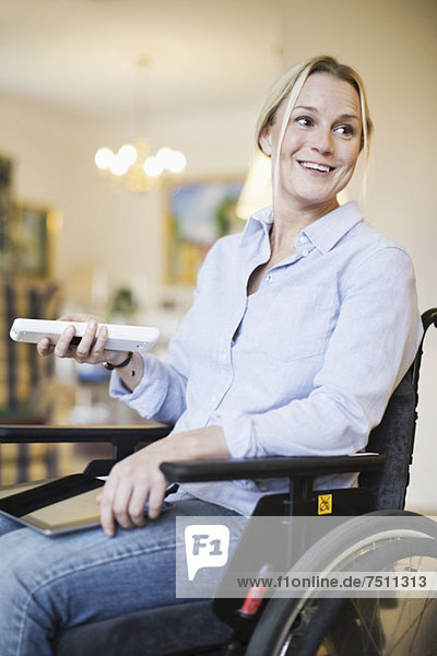 Happy disabled woman in wheelchair with digital tablet looking away while using remote control