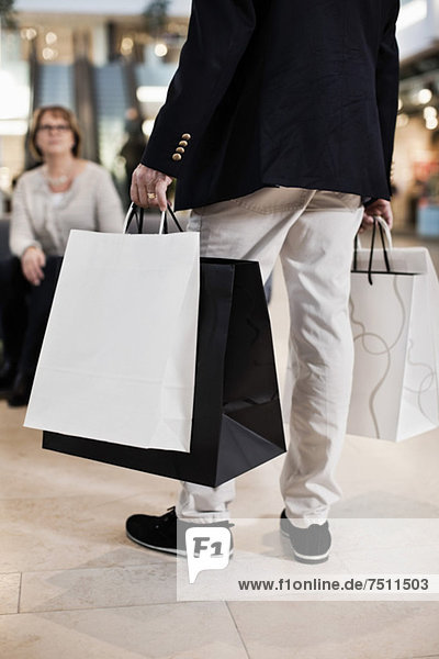 Low section of man carrying shopping bags with woman in background at mall