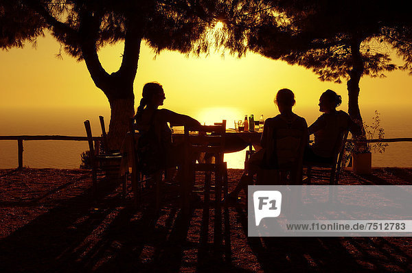 Drei Frauen sitzen bei untergehender Sonne an einem Tisch  Abendstimmung in einem griechischen Garten am Meer  Lefkas  Griechenland  Europa