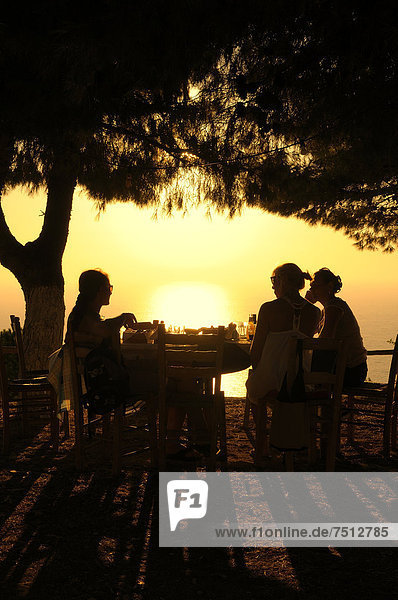 Drei Frauen sitzen bei untergehender Sonne an einem Tisch  Abendstimmung in einem griechischen Garten am Meer  Lefkas  Griechenland  Europa