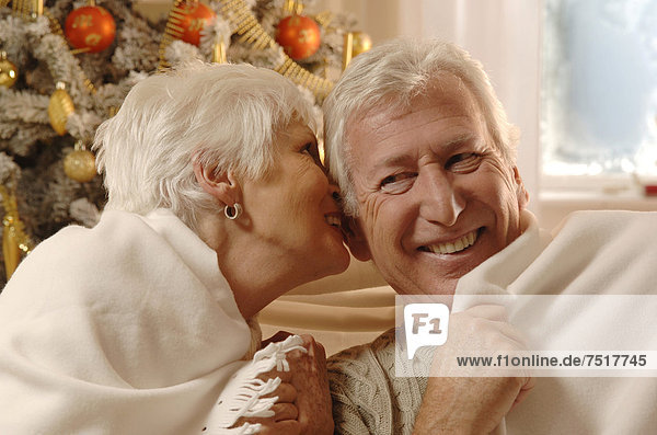 Älteres Pärchen sitzt verliebt mit Decke vor dem Weihnachtsbaum