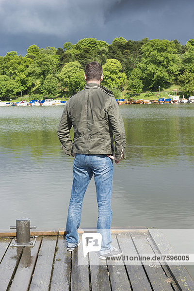 Mann steht auf einem Bootssteg an einem See und schaut auf die dunklen Regenwolken