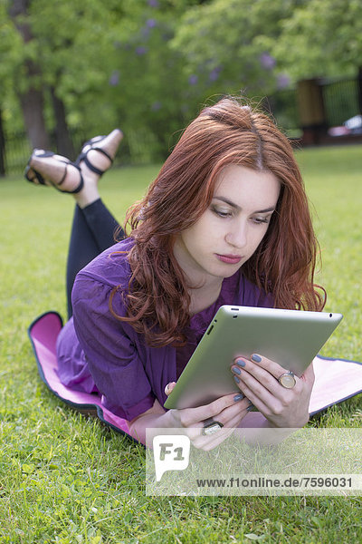 Junge Frau liegt in einem Park auf dem Rasen und liest etwas auf einem Tablet-PC