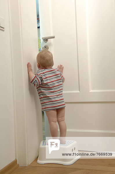 Kleinkind steht auf Hocker  schaut durch Türspalt