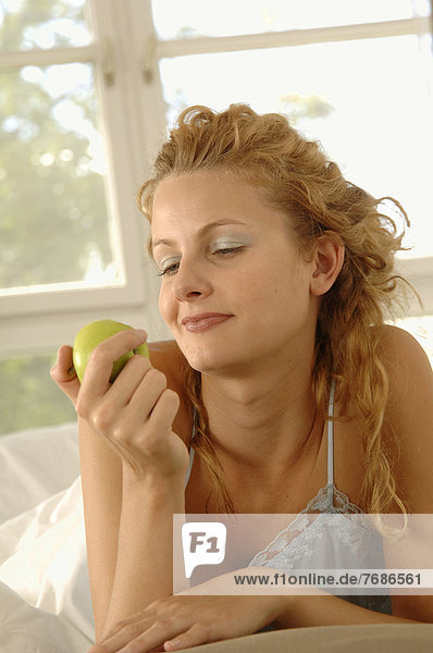 Blonde Frau  25-35 Jahre  liegt aufgestützt auf Bauch im Bett mit Apfel in der Hand