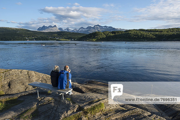 Couple sitting on a bench looking out to the tidal Saltstraumen strait in the evening light  Børvasstindan mountain range at back