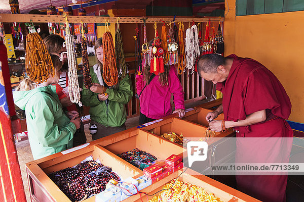 monk works on some beaded jewelry at Drepung Monastery  Tourists look at jewelry as  Lhasa  Tibet  Xizang  China