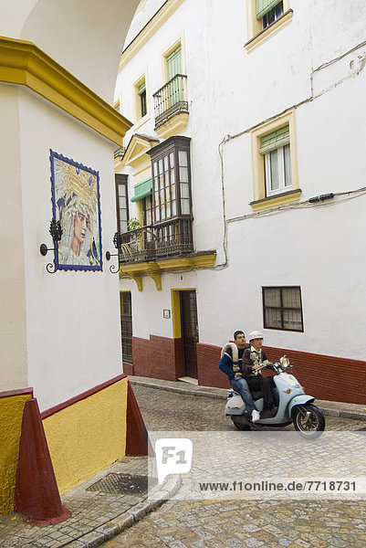 Two Young Men On Moped Passing Virgin Mary Ceramic On House Wall In Alley Of Jerez  Andalucia  Spain