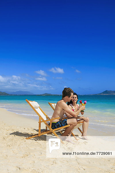 Hawaii  Young Couple On The Beach In Lounge Chairs With Tropical Drinks.