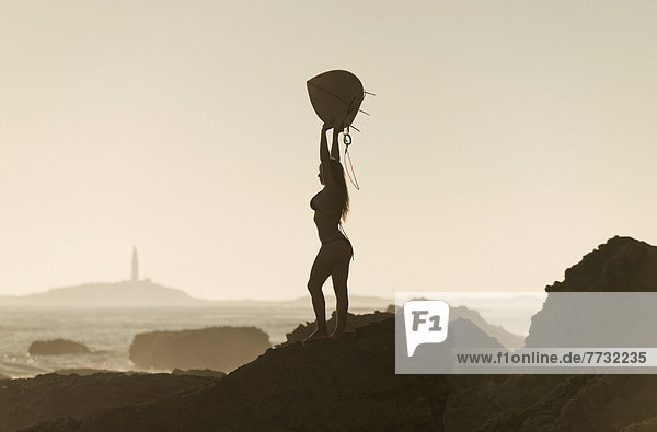 Silhouette of a female surfer holding her surfboard up above her head while standing on a rock at the coast  andalusia spain