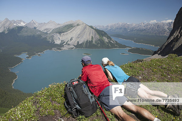 Male And Female Hiker Laying Down On A Mountain Ridge Overlooking An Emerald Lake And Mountains Below With Blue Sky In Kananaskis Provincial Park  Alberta Canada