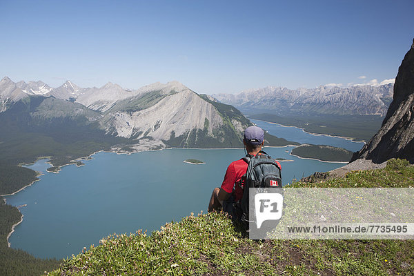 Male Hiker Sitting On A Mountain Ridge Overlooking An Emerald Lake And Mountains Below With Blue Sky In Kananaskis Provincial Park  Alberta Canada