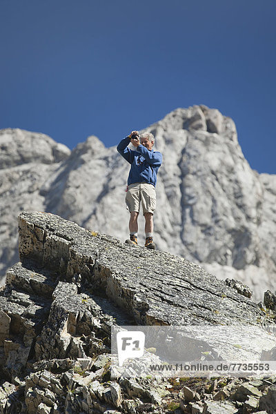 Male Hiker On Top Of Rock With A Camera Taking Photos With Mountain And Blue Sky In Background In Kananaskis Provincial Park  Alberta Canada