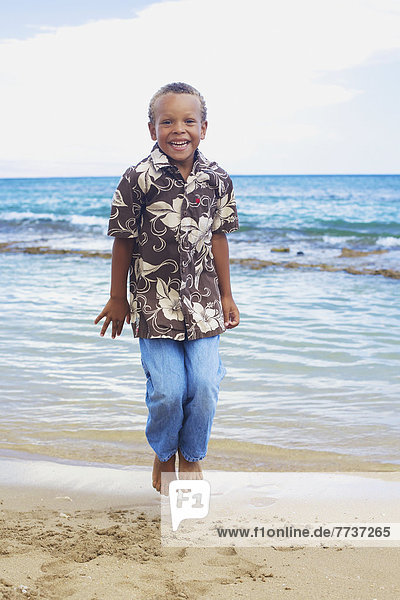 A young boy jumps with joy on the sand at the water's edge Hawaii united states of america