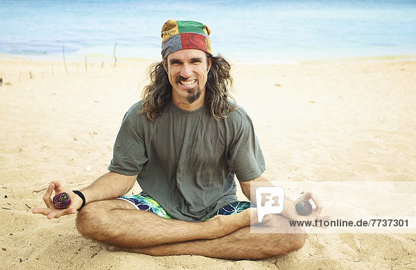 A man sits in the sand in a yoga pose holding two hackey sacks Hawaii united states of america