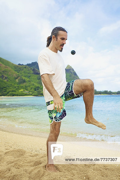A man bumps his hackey sack with his knee while standing in the sand at the water's edge Hawaii united states of america
