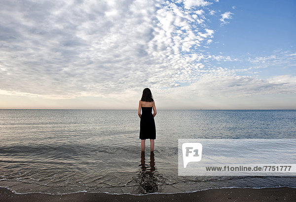 Woman wearing a black dress standing in the water of the sea  moody clouds