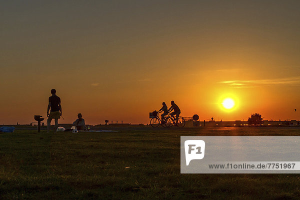 Ein Paar grillt auf dem Tempelhofer Feld in der Abendsonne  hinter ihnen fahren zwei Radfahrer