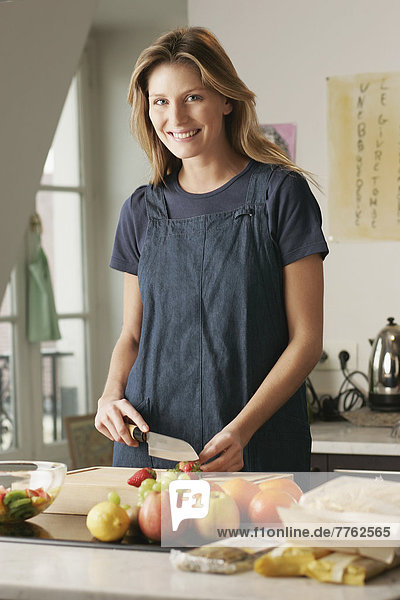 Smiling young woman in apron cutting strawberries on chopping board  posing for the camera  apples  oranges  lemon  white grapes and salad bowl on table
