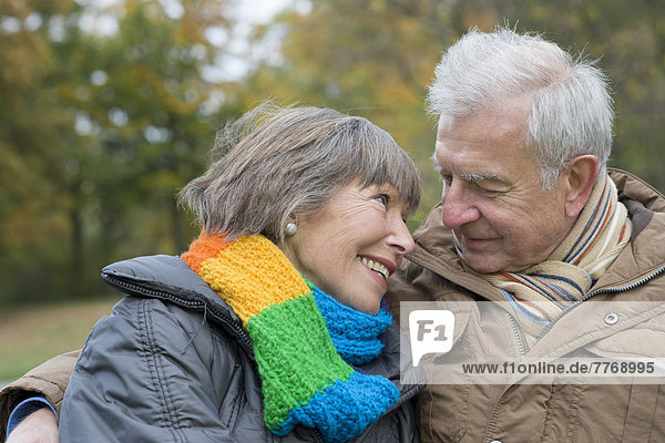 Seniorenpaar sitzt Arm in Arm auf Parkbank und lächelt sich an