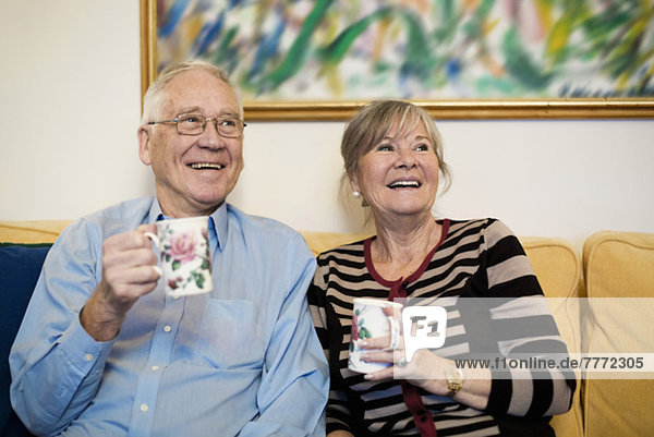 Glückliches Senior-Paar schaut weg und hält Kaffeetassen auf dem Sofa.