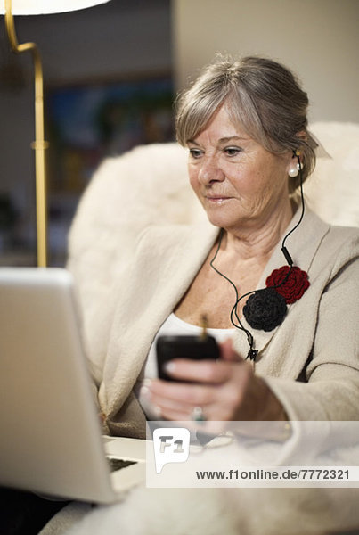 Senior woman using laptop while listening music through mobile phone on couch
