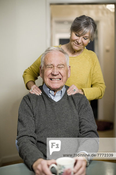 Älterer Mann  der die Zähne zusammendrückt  während die Frau ihm zu Hause eine Schultermassage gibt.