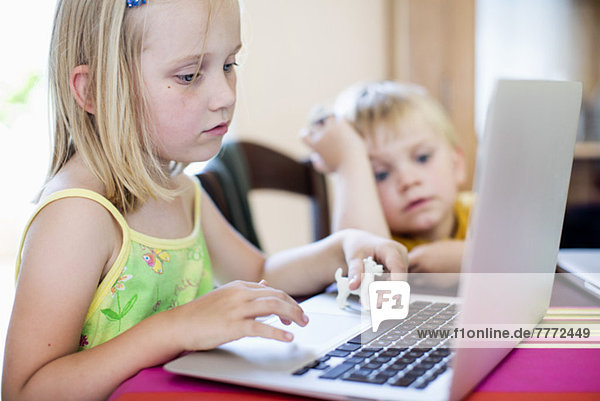 Girl using laptop at table with brother sitting in background