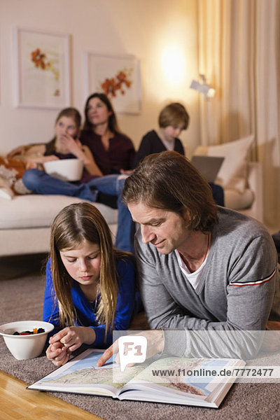 Father and daughter reading map on floor with family sitting on sofa in background