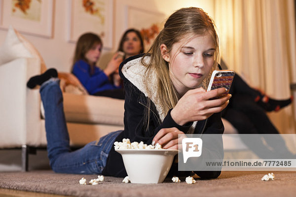 Mädchen mit Handy und Popcorn auf dem Boden mit Familie im Hintergrund