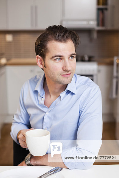 Mittlerer erwachsener Mann mit Kaffeetasse  der beim Sitzen am Frühstückstisch wegschaut.