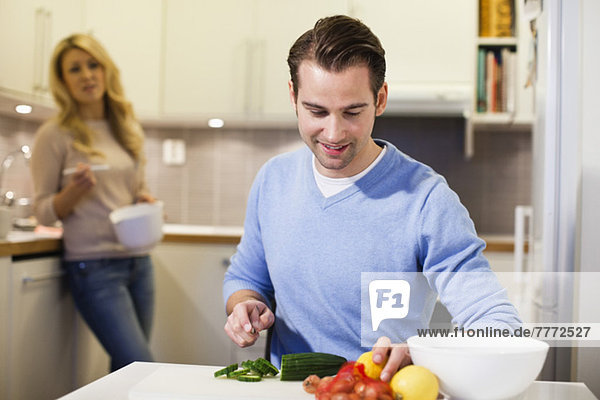 Mittlerer Erwachsener Mann beim Gurkenhacken an der Küchentheke mit Frau im Hintergrund