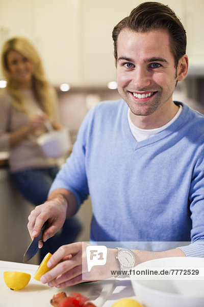 Porträt eines erwachsenen Mannes  der Zitronen an der Küchentheke mit Frau im Hintergrund schneidet.